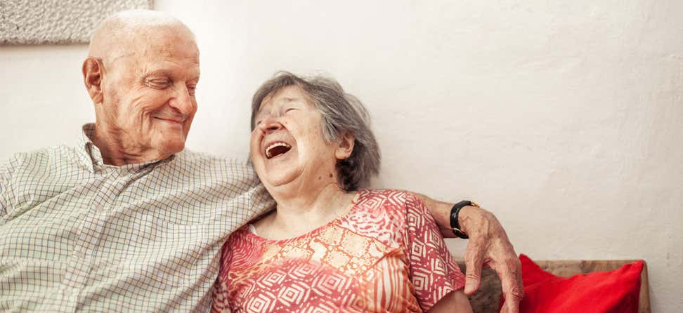 A senior couple sit on a couch, laughing and enjoying each other's company.