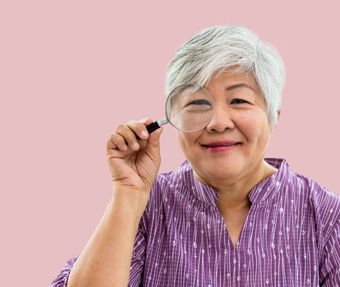 A senior Asian woman smiles while holding a magnifying glass to her face. 