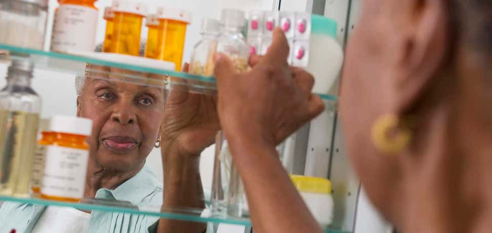 An older black women is reaching for a prescription bottle in her medicine cabinet. 