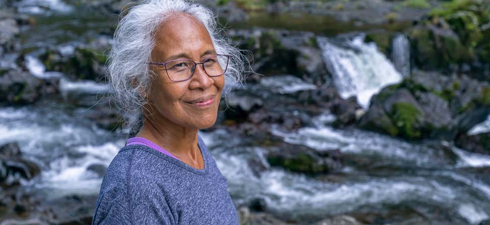 A senior Pacific Islander woman is standing near a river, smiling as she takes a walk in nature.