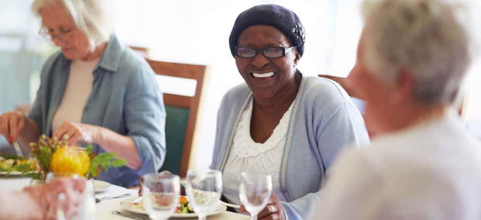 A senior Black lady wearing glasses laughs with her friends during lunchtime at a senior center.