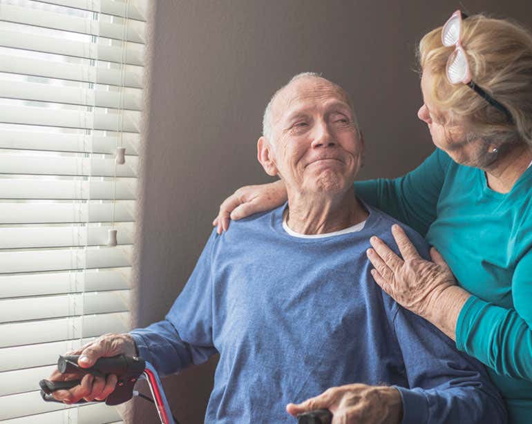 An older caregiver is seen at home helping a male senior - both looking at each other. 