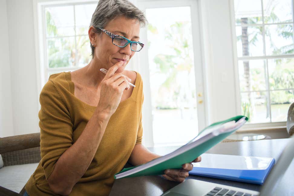 An older woman with glasses looks at paperwork and her laptop with a pen in her hand