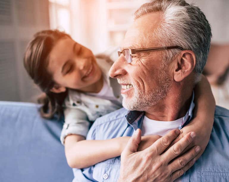 A grandfather with a beard and glasses is being hugged by his granddaughter while he's sitting on the couch. Both are smiling.