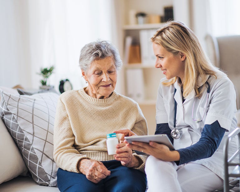 A Caucasian female nurse, caregiver is giving instructions to a Caucasian female senior who's sitting on the couch.