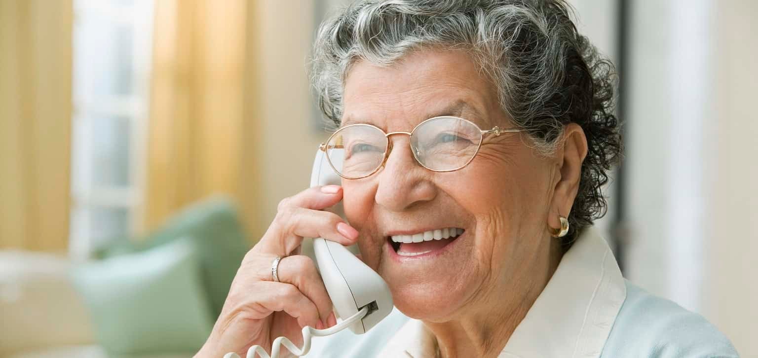 An older woman is talking on her landline, smiling and looking off camera.