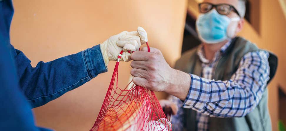 A senior male wearing a mask is receiving a grocery delivery from a caregiver wearing gloves during the pandemic.