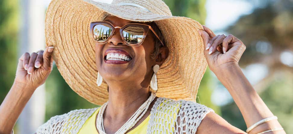 A Black senior woman is radiant in the sun, wearing sunglasses and a brimmed hat while smiling.