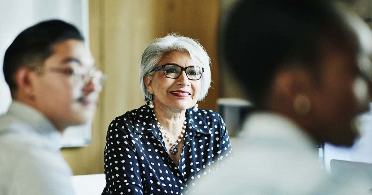 A senior female with glasses on sits in a room of professionals during a meeting.