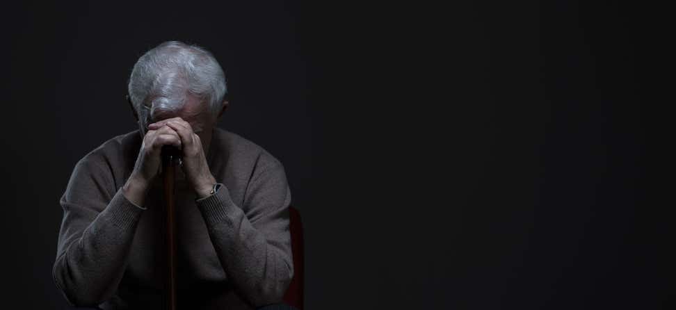 A Caucasian senior man is sitting in the dark with his head bent over on his cane, seemingly depressed.