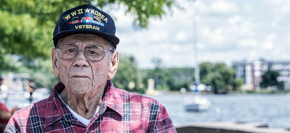 A close up shot of a senior male Veteran looking straight at the camera with the beach/harbor in the background.