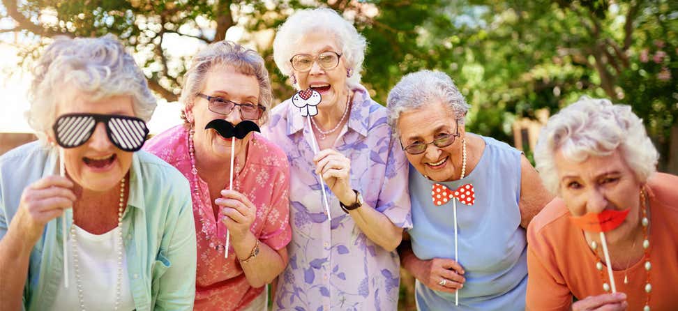 A group of senior women are having fun, holding up mustache/various masks while posing for a photo together.
