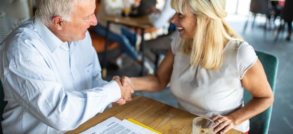 A senior Caucasian female shakes hands with a senior Caucasian man. Both are sitting at a table, discussing a financial agreement.