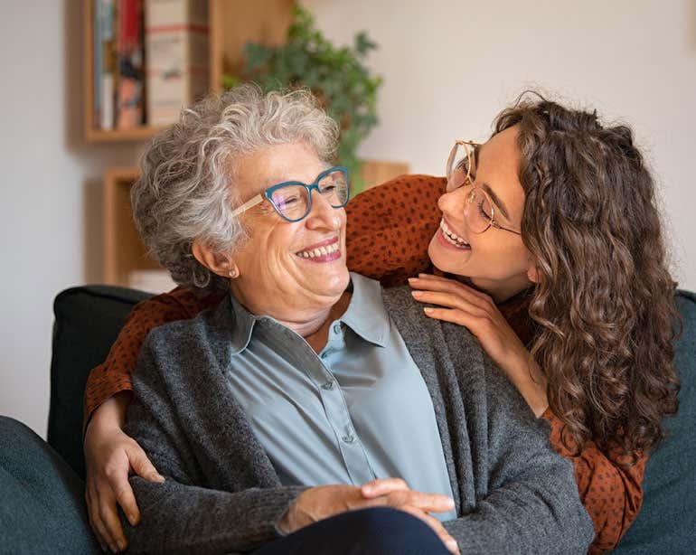 A senior woman wearing glasses is smiling while being hugged by her younger female caregiver, that's also wearing glasses.