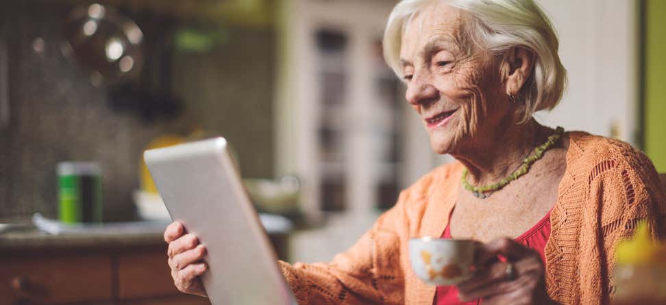 A senior woman uses her tablet to calculate her finances while drinking hot tea.