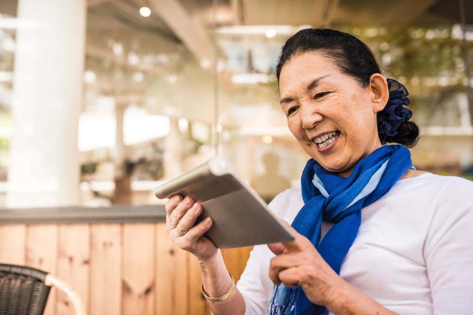 An older Asian woman is looking at her tablet while seated outside at a cafe.
