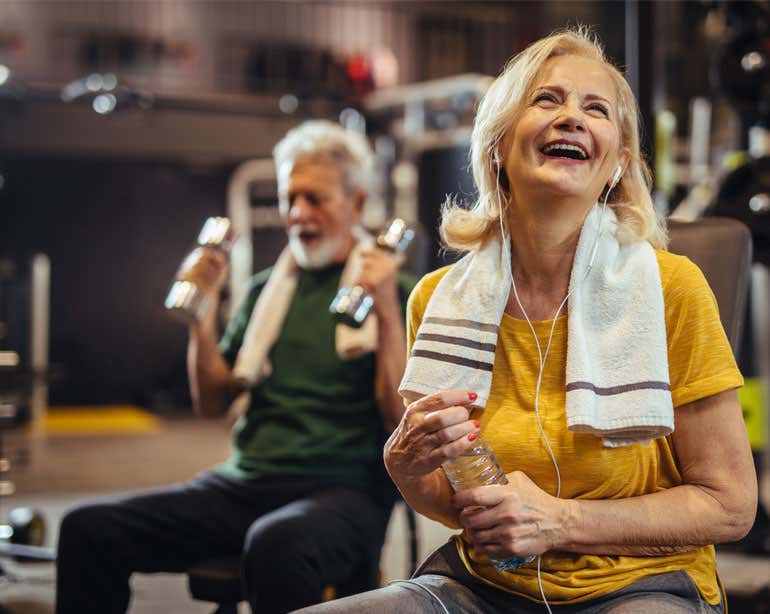 A senior woman in her gym clothes takes a break from exercising while smiling and holding a bottle of water. Her husband is in the foreground seen lifting dumbbells.