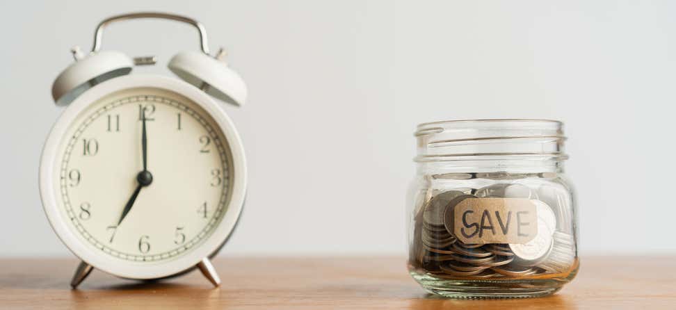 An old alarm clock sitting to the left of a mason jar labeled "save", indicating a race for time to save money.