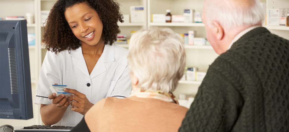 A Black female pharmacist is helping an older senior couple, explaining a medication to them as they check out.