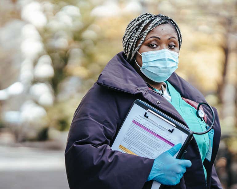 A Black healthcare professional is standing outside with a mask and gloves on, holding a clipboard.