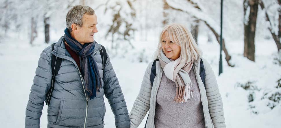 A senior couple is walking hand-in-hand outside in the winter, where the foreground is covered in snow.