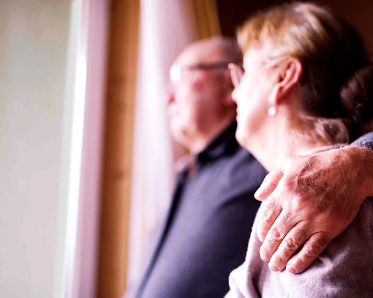 A senior couple embraces each other, staring out of their window.