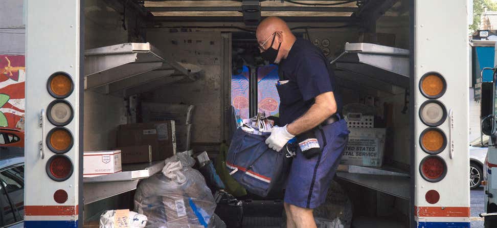 A masked postal worker in New York City holds a canvas bag full of mail and is loading it into his vehicle before heading out for the day.