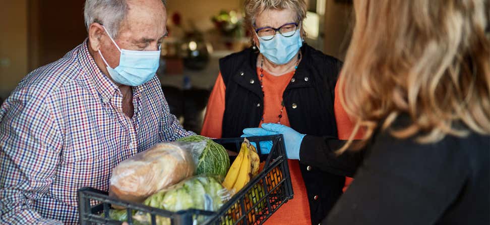 A professional from a community-based organization is delivering a plastic cart full of fresh produce to an elderly couple during the pandemic.