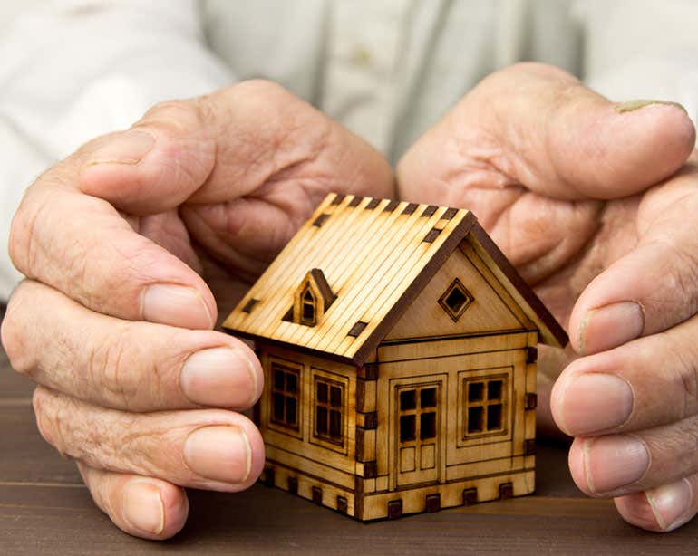 Senior hands are seen cupping a small wooden house, indicating that they're protecting their investment.