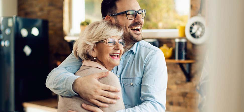 A senior Caucasian woman is seen embracing her younger male caregiver, both are smiling.