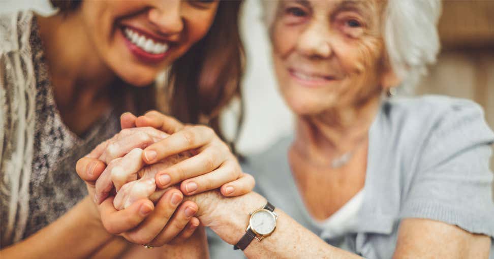 A younger female caregiver holds the hands of a senior Caucasian woman who is smiling.