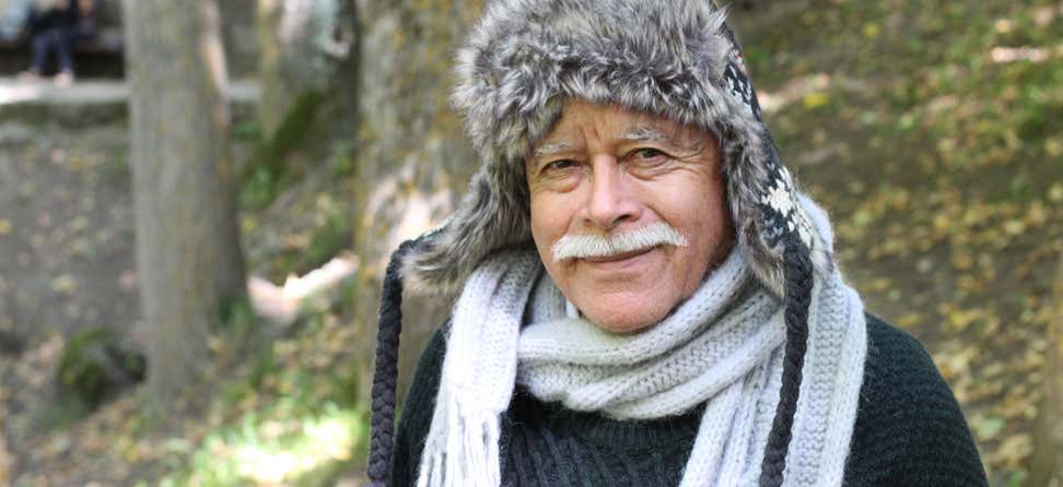 A senior eskimo man, wearing a warm hat and scarf, is looking at the camera smiling. 
