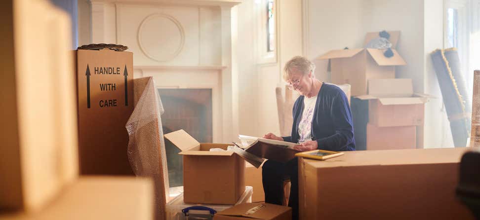 A senior woman is sitting in her house, full of boxes that are packed - looking at a photo album and remembering the past.