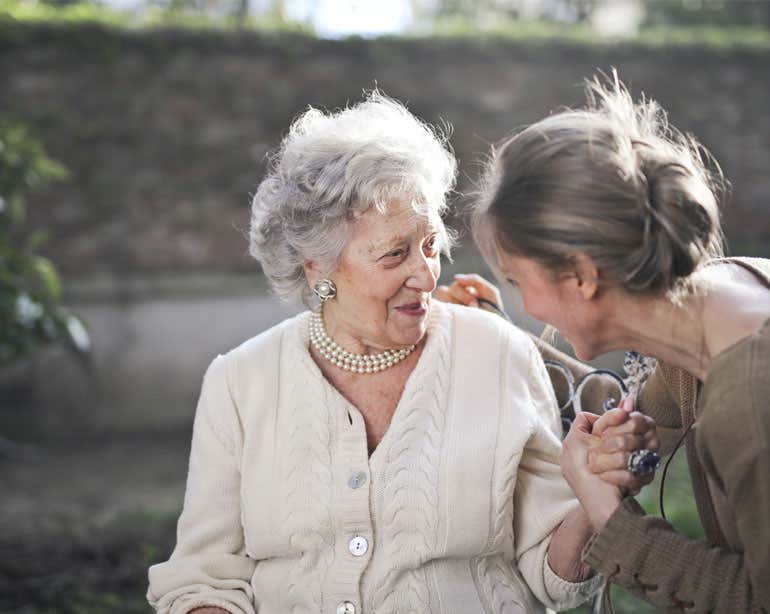 An older female caregiver is seen holding the hand of a Caucasian senior woman. Both are smiling.