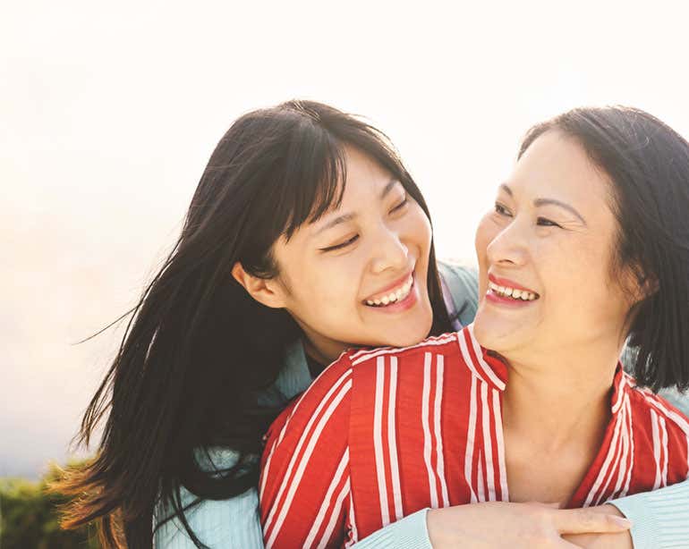 A young female Asian caregiver is embracing her senior mom outside, both are laughing.