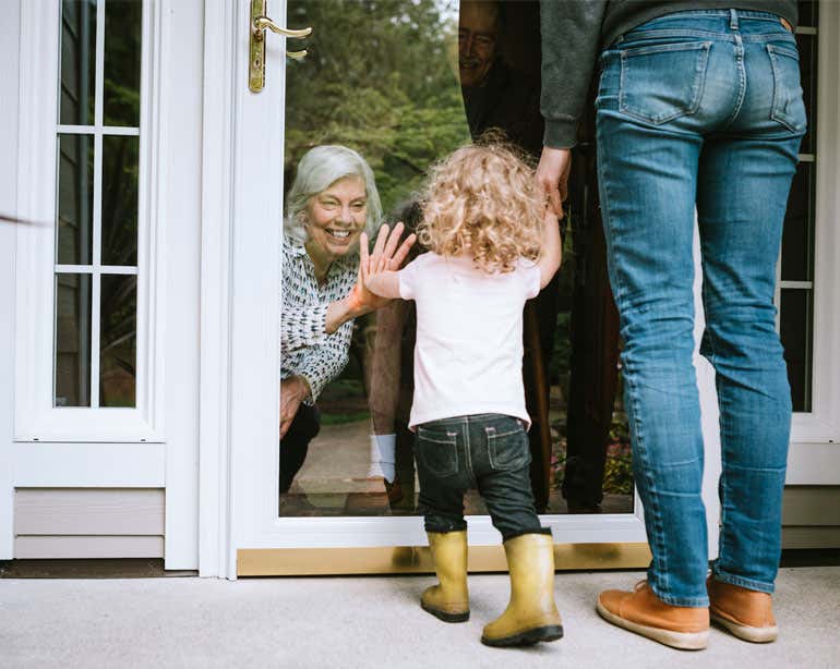 A grandmother is waving to her granddaughter and daughter through a sliding glass window, being careful to practice social distancing during the pandemic.
