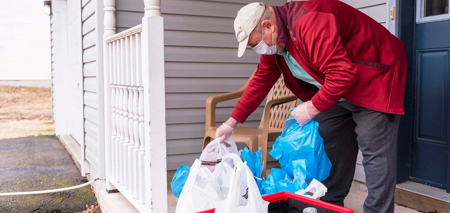 Senior man wearing a protective mask and gloves is taking groceries out of the bins left on his porch into his house during COVID-19 pandemic outbreak.