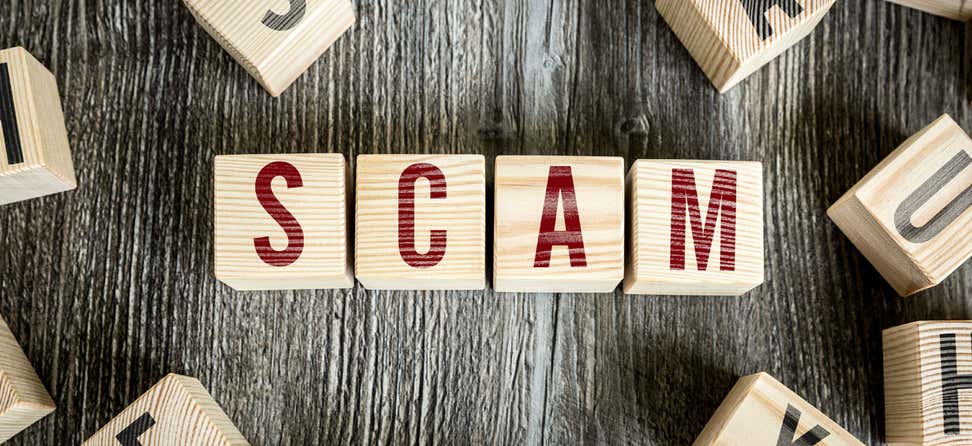 The word "SCAM" is spelled out in wooden blocks on an old picnic table. 