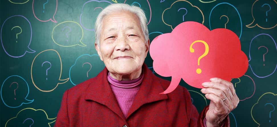 An older Asian woman is holding a red thought bubble sign in her hand that has a yellow question mark inside of it.