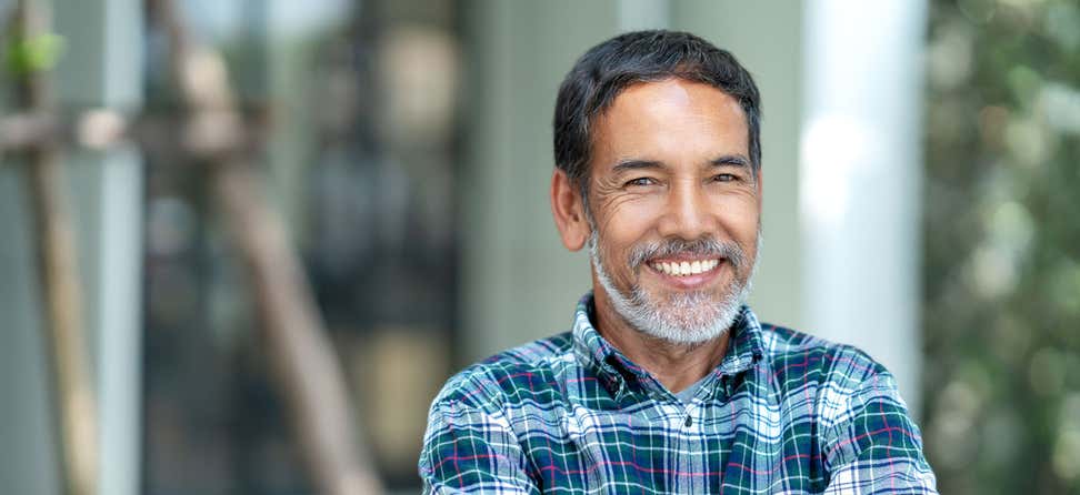 A Hispanic senior man wearing a blue-green plaid shirt is standing outside smiling.