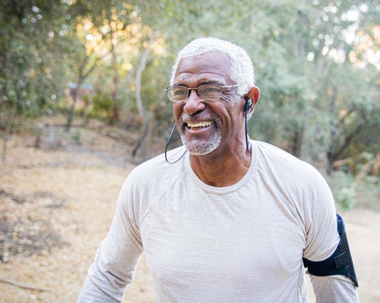 A Black senior man is exercising outside, wearing his phone on his arm and with his earbuds in.