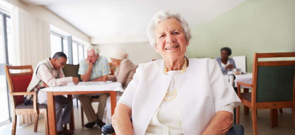 An older woman smiles while sitting in a room with other seniors at a senior center.