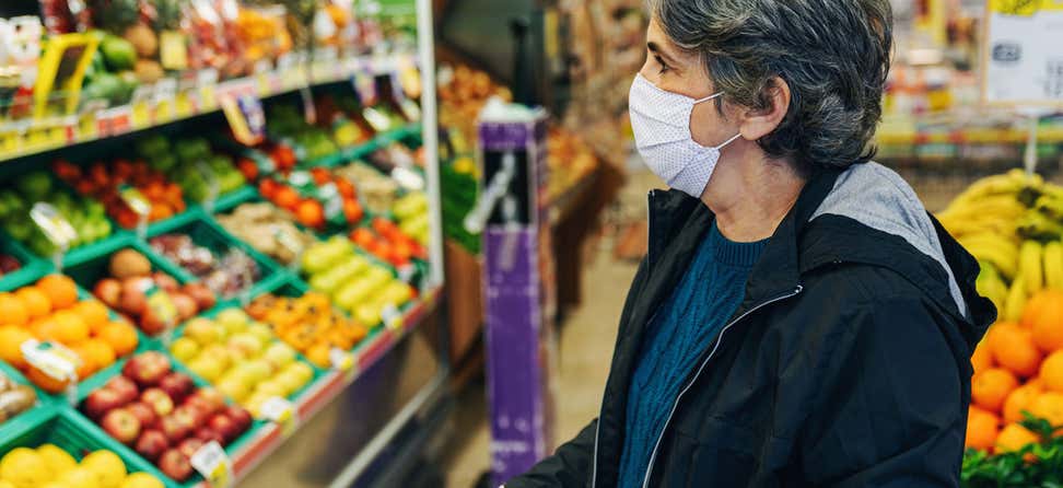 A senior woman with dark, short hair and wearing a mask is in the produce section of a grocery story contemplating what to buy.