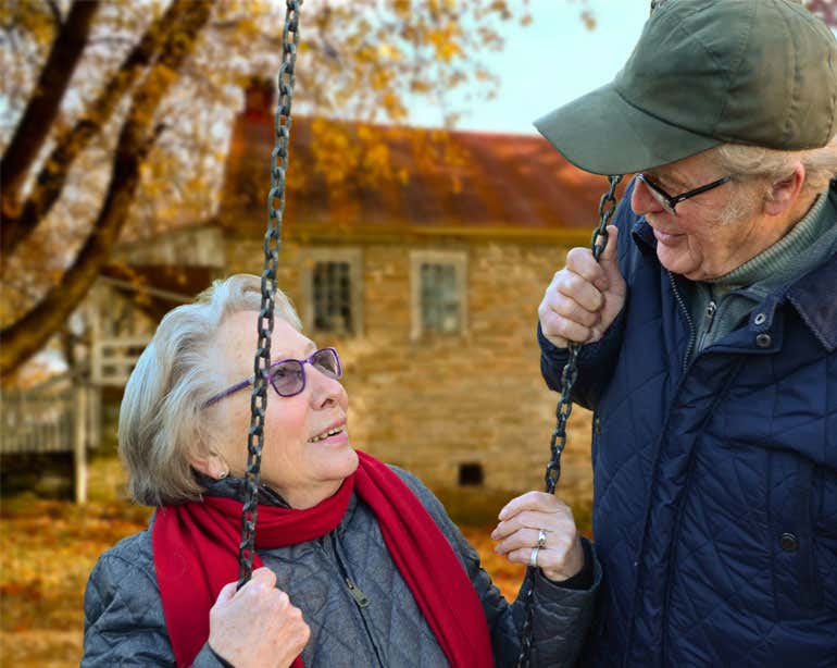 A senior Caucasian man looks down lovingly at his wife while she's swinging outside.
