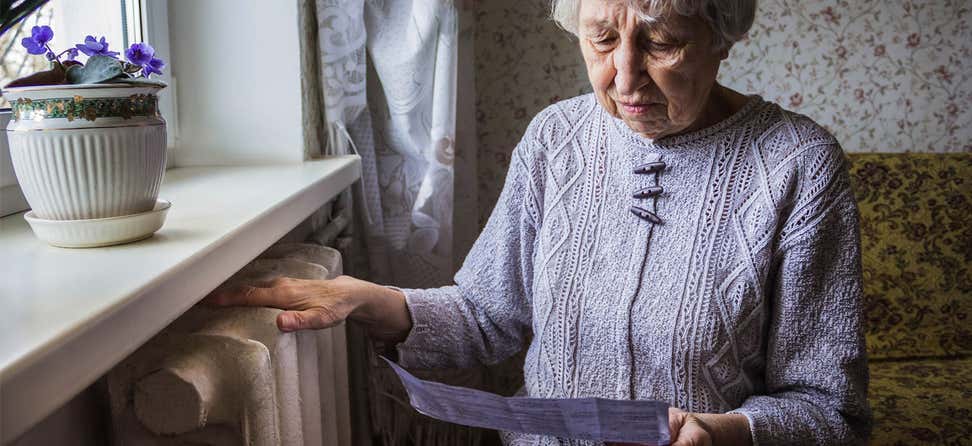 A senior woman places her hand on her heater/radiator while looking at her electricity bill.