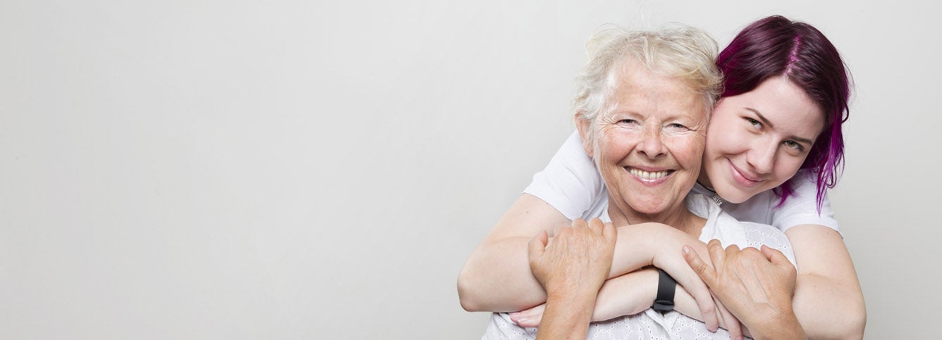 A senior woman is smiling while her younger female caregiver embraces her with a hug from behind.