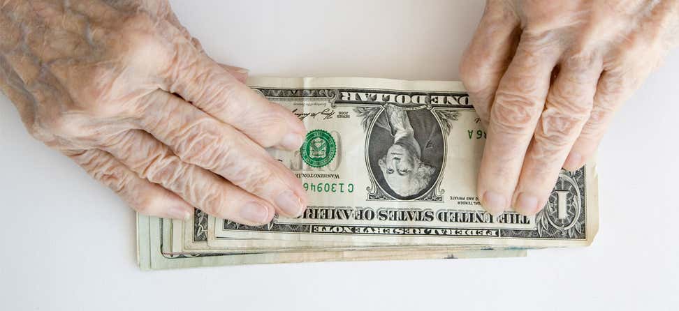 Older/senior hands are placed on top of a stack of one dollar bills.
