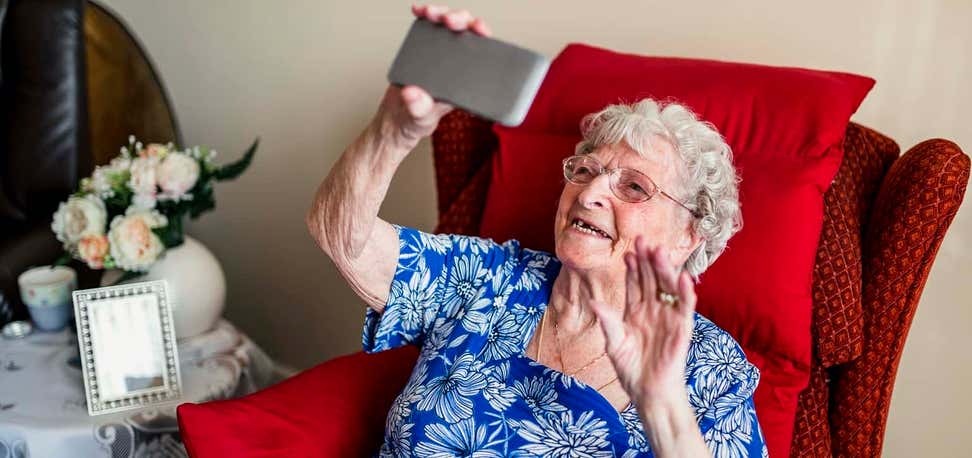 An older woman is holding her mobile phone up, having a video call with a family member.