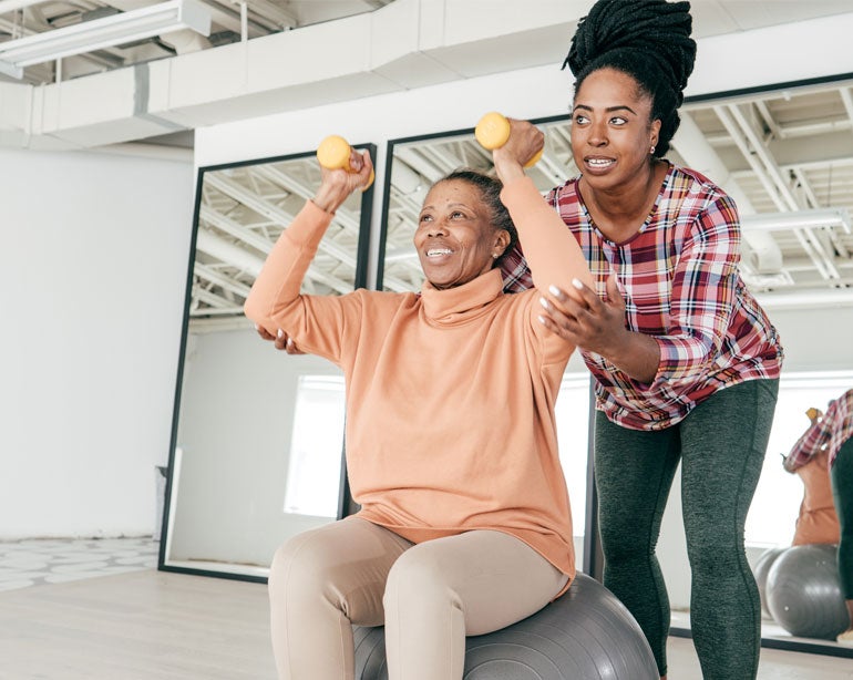 Black woman sitting on exercise ball in gym with hand weights and physical therapist helping her