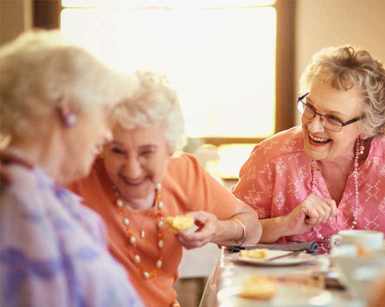 Three senior Caucasian women are laughing while enjoying a meal at a senior center.